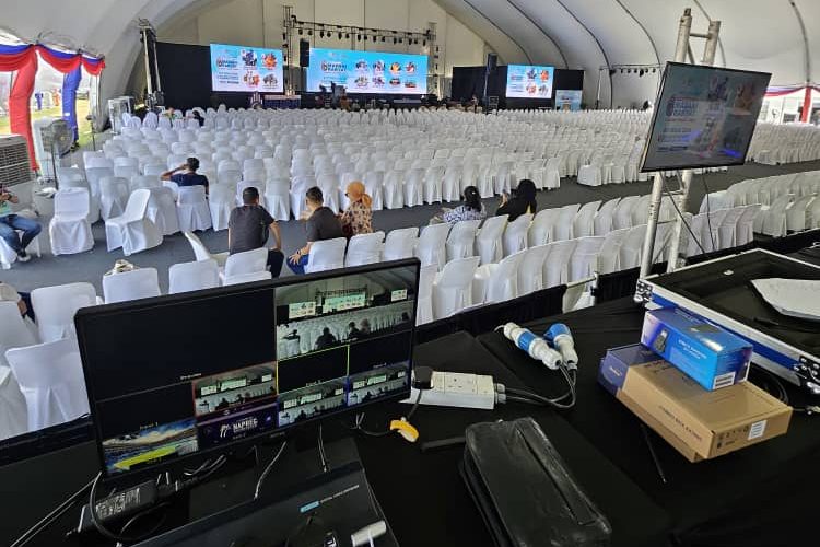 A view from an event control booth showing video production by Zaramiz Sdn Bhd monitoring screens overlooking a large empty indoor hall filled with rows of white chairs.