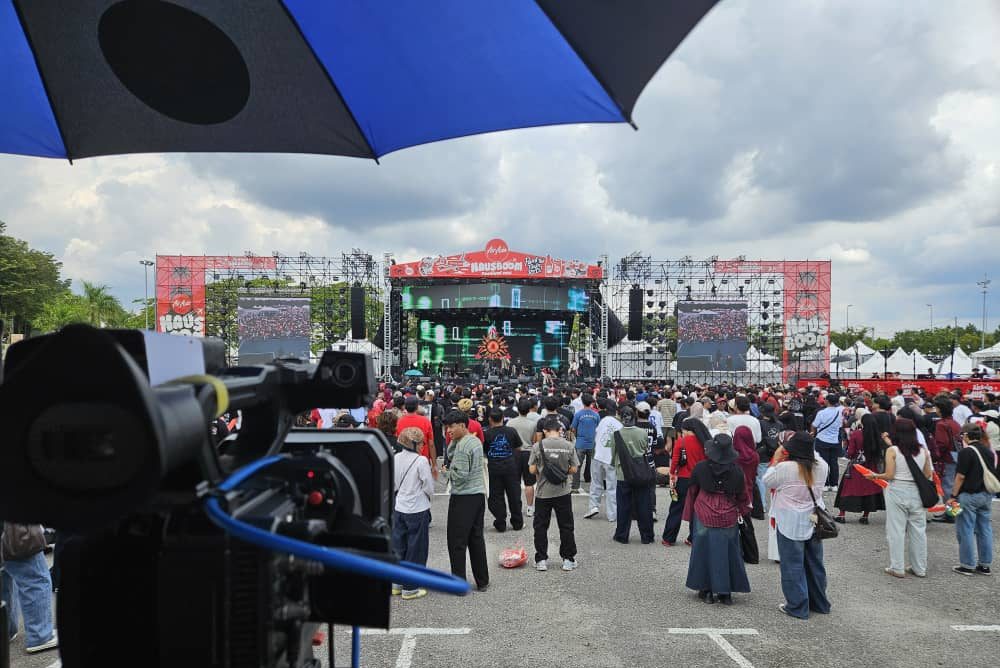 A large professional production camera owned by Zaramiz Sdn Bhd facing an outdoor crowd at a large public event or concert stage under an umbrella.