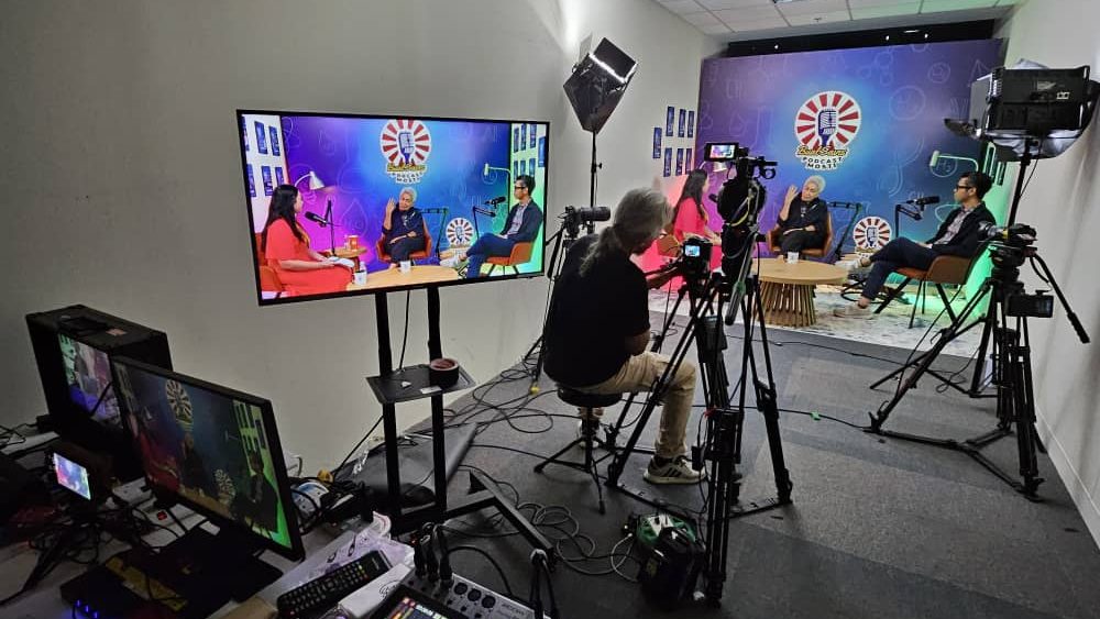 Behind-the-scenes view of a professional multi-camera talk show setup by Zaramiz Sdn Bhd, showing crew members operating video cameras, monitoring screens, studio lighting, and guests recording an interview on a stage.