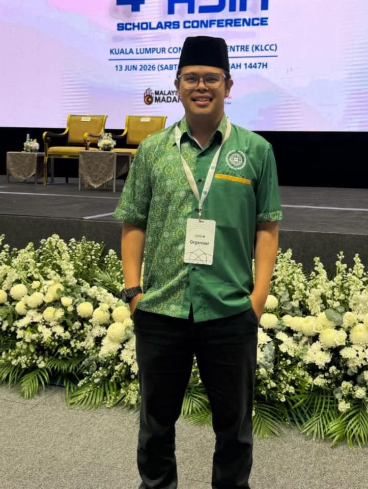 Profile photo of Huzairy Osman, Production Executive at Zaramiz Sdn Bhd, standing in a green polo shirt in front of an event stage setup.