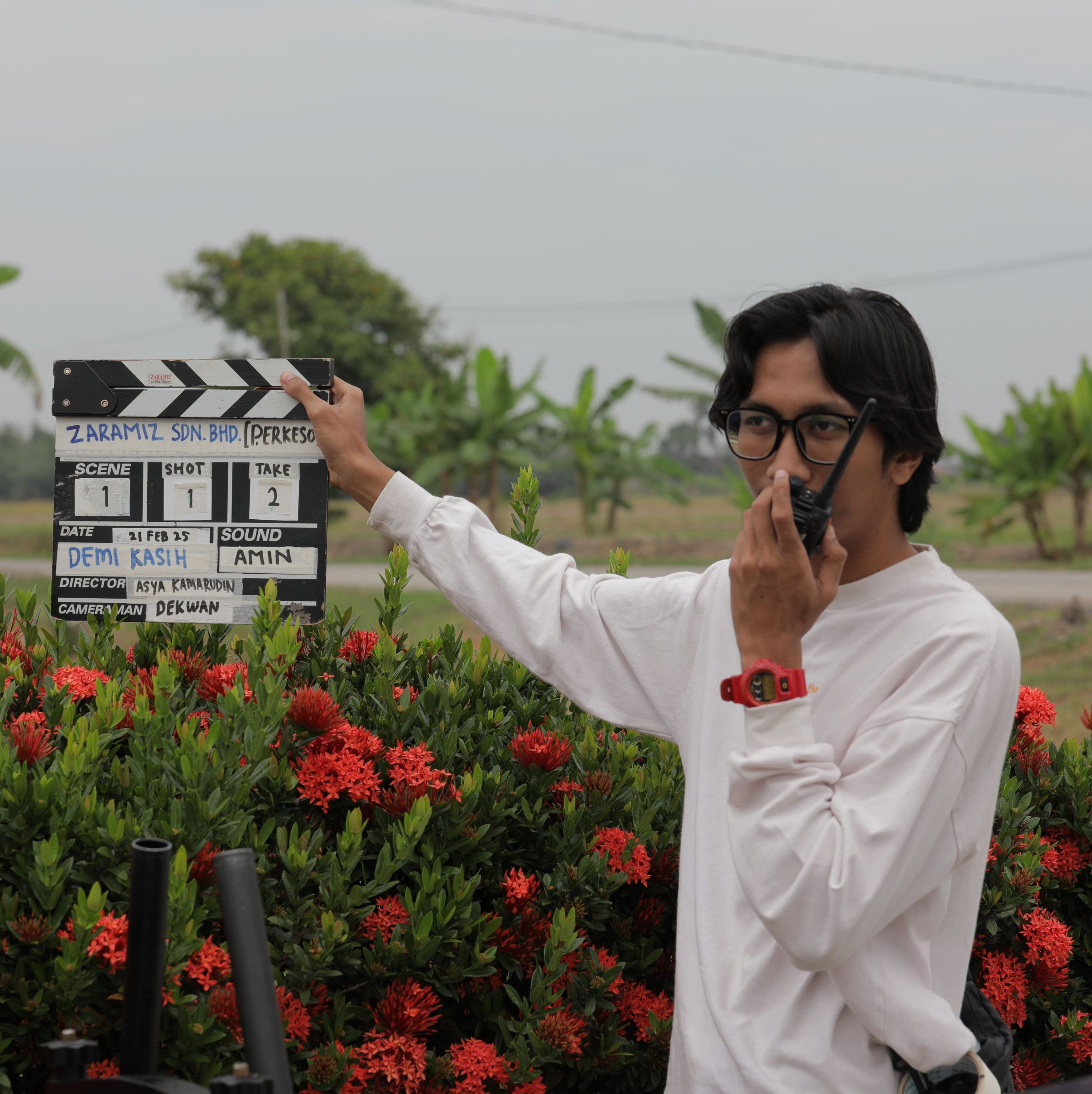 A crew member from Zaramiz Sdn Bhd holding a film clapperboard and communicating via walkie-talkie on an outdoor production set.