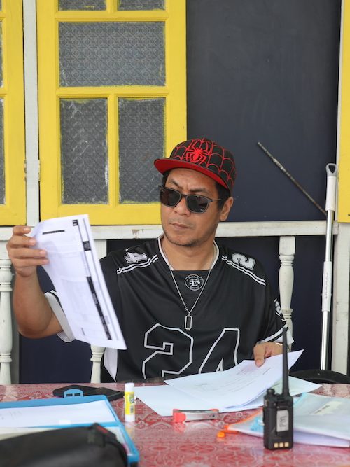 Profile photo of Es Hairee, Production Manager at Zaramiz Sdn Bhd, wearing sunglasses and holding production paperwork outdoors.
